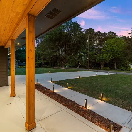 Covered wooden porch with recessed lighting overlooking a lit concrete driveway and green yard at dusk.