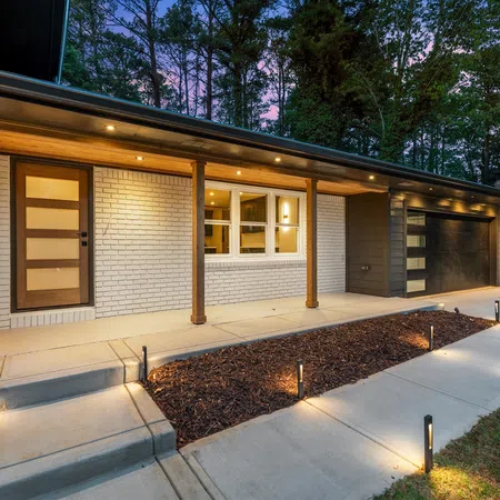 Modern house exterior with white brick, wooden door, garage, porch lights, and landscaped pathway at dusk.