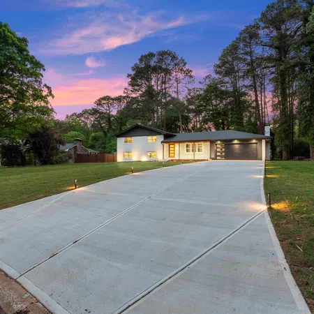 Modern house with a long concrete driveway, illuminated pathway lights, and a sunset sky in a wooded neighborhood.