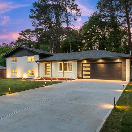 Modern split-level home with illuminated driveway and garage during dusk surrounded by tall trees.