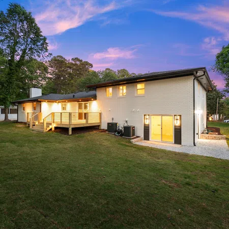 Modern two-story white brick house with wooden deck, large backyard, and glowing interior lights at sunset.