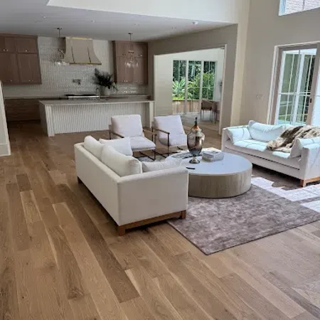 Spacious modern living room with white sofas, armchairs, wooden flooring, and an open kitchen in the background.
