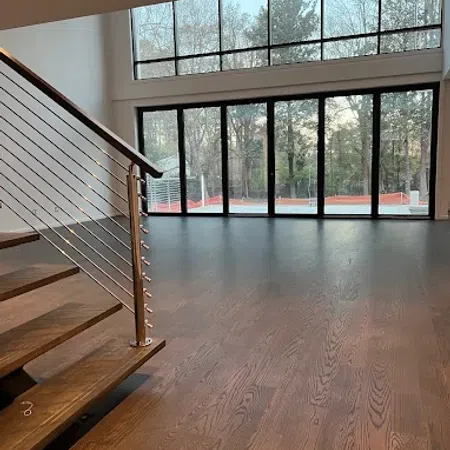 Modern interior with wooden flooring, metal railing staircase, and large floor-to-ceiling windows showing a forest view.