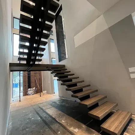 Modern floating wooden stairs with black metal supports in a spacious, unfinished interior with tall windows.