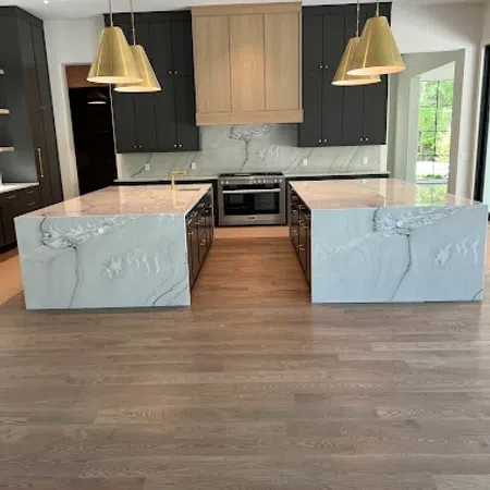 Modern kitchen with dual marble islands, black cabinets, wood floors, and gold pendant lights above.