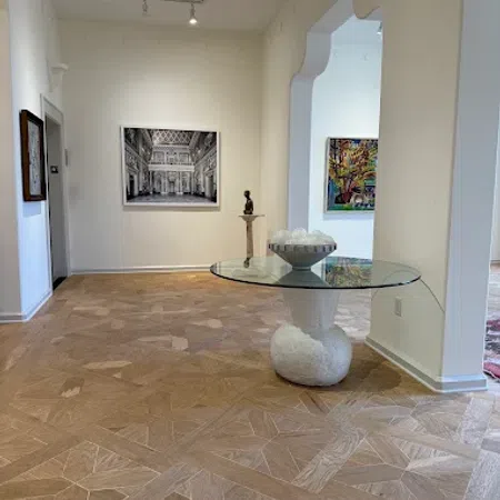 Modern art gallery room with geometric wooden floor, glass table, sculpture, and framed paintings on white walls