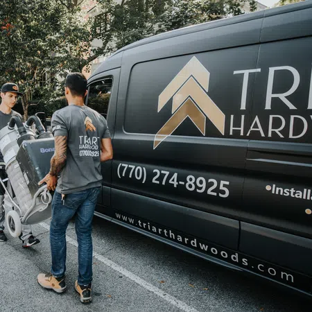 Two workers unloading equipment from a Tri Art Hardwood van in an outdoor setting with trees and pavement.