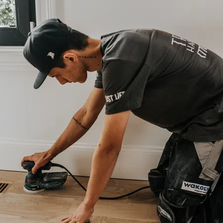 Man in cap sanding wooden floor near white wall using electric sander powered by Bosch