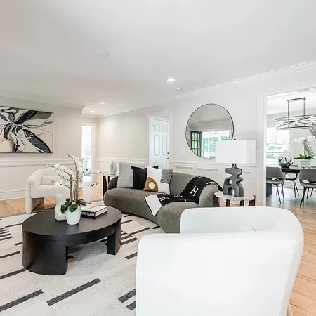 Bright modern living room with gray sofa, white armchairs, round black coffee table, and abstract wall art.