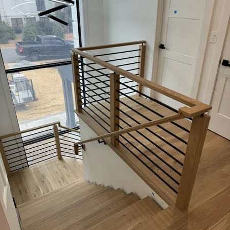Modern staircase with wooden steps and black metal railing next to large window in bright interior.