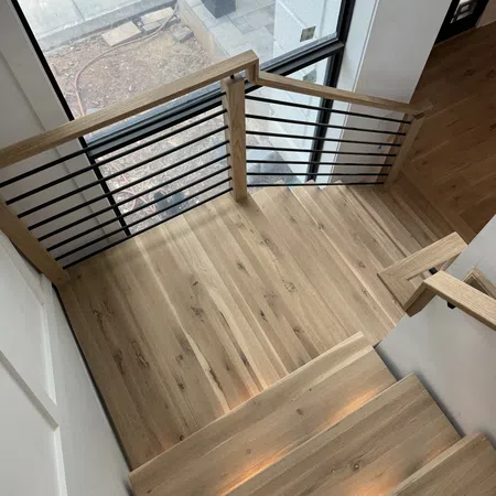 Top view of modern wooden staircase with black metal railings and large window overlooking outdoor area.