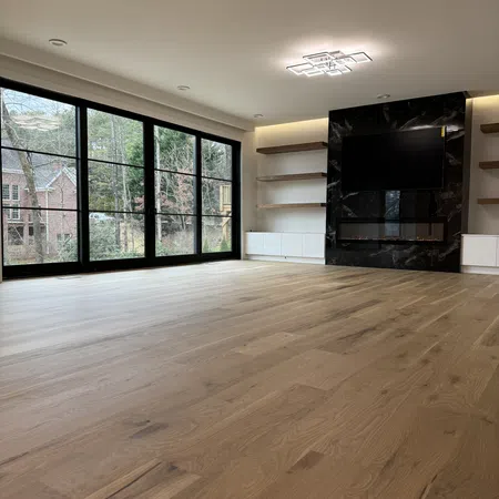 Modern empty living room with light wood flooring, large black-framed windows, wall-mounted TV, and fireplace.