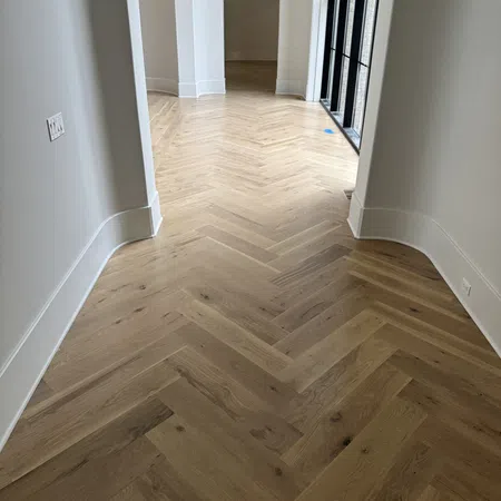 Empty hallway with light herringbone wood flooring, white walls, curved baseboards, and arched doorway.