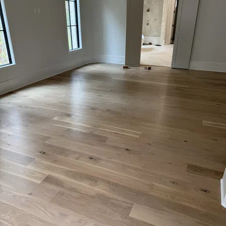 Empty room with light hardwood flooring, white walls, large windows, and an open doorway leading to a tiled area.