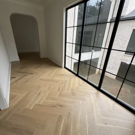 Empty room with light wood herringbone flooring and large black-framed floor-to-ceiling windows