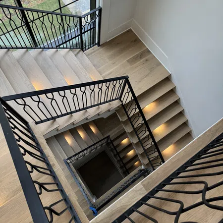 Top-down view of a modern wooden staircase with black metal railings and warm step lighting in a bright interior.