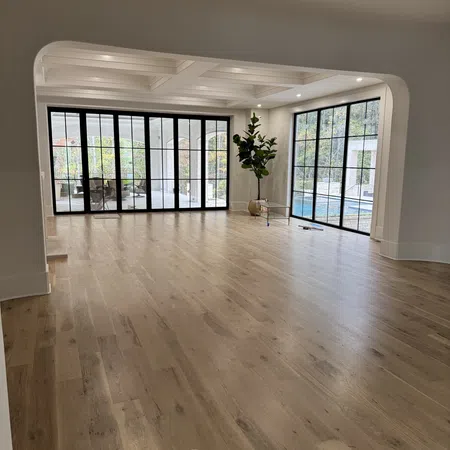 Spacious empty room with light hardwood floor, large black-framed windows, coffered ceiling, and indoor plant.