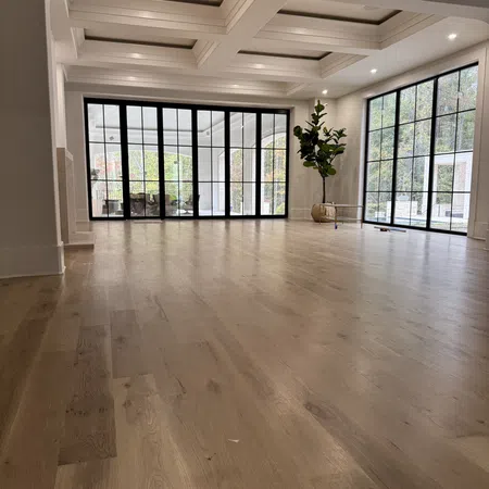 Spacious room with light wood flooring, large black-framed windows, coffered ceiling, and a potted plant.