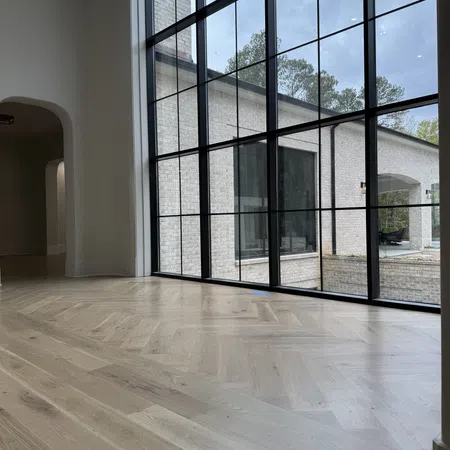 Modern empty room with large black-framed floor-to-ceiling windows and light wood parquet flooring.