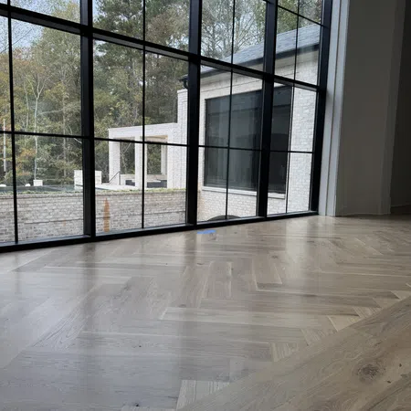 Large modern window with black grid frames overlooking trees and a white brick building, interior herringbone wood floor.