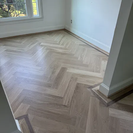 Light wood herringbone pattern flooring with dark wood border in an empty room with white walls and large window.