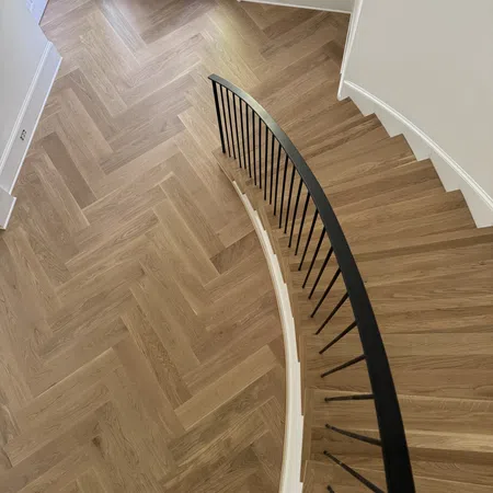 Curved wooden staircase with black railing and chevron-patterned wood flooring in entryway with dark door.
