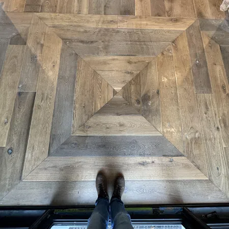 Top-down view of feet standing on a geometric patterned wooden floor with natural wood tones and textures.