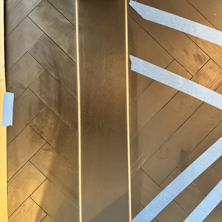 Dark wood herringbone flooring with blue painter's tape marking sections near white trim and a central plank.