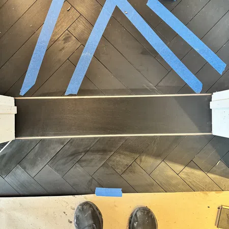 Dark wood herringbone floor with blue painter's tape and feet in work boots on a construction site.