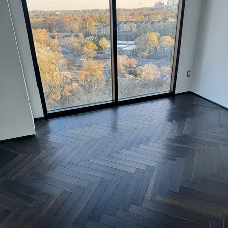 Dark herringbone wood floor in empty room with large window overlooking autumn cityscape.