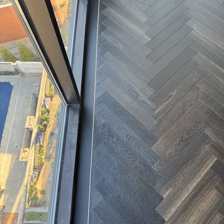 Dark herringbone wood flooring next to a tall window overlooking an urban street with plants and buildings.