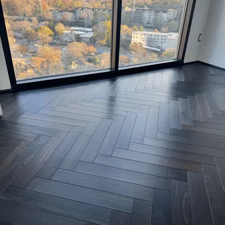 Dark herringbone hardwood floor in modern room with large window showing city skyline and autumn trees.