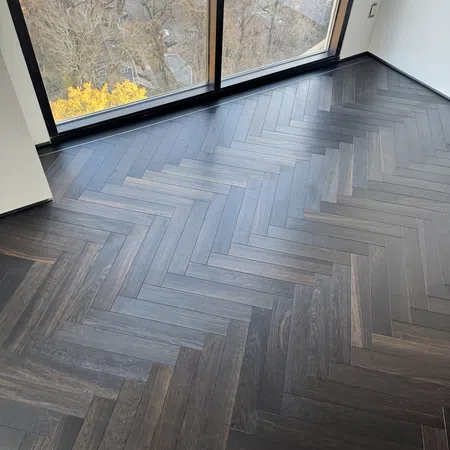 Modern room with large floor-to-ceiling window and dark herringbone wood flooring overlooking autumn trees.