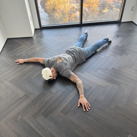 Man lying face down on dark herringbone wood floor in bright empty room with large window view.