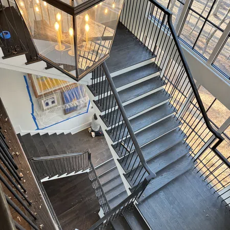 Top view of modern dark wood staircase with black metal railing and large window lighting the space.