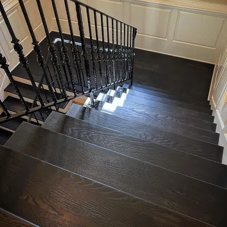 Dark wooden staircase with decorative black metal railing and white paneled wall in bright interior.