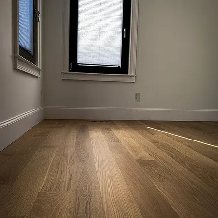 Empty room with wooden flooring, white walls, and two windows with blinds partially closed letting in sunlight.