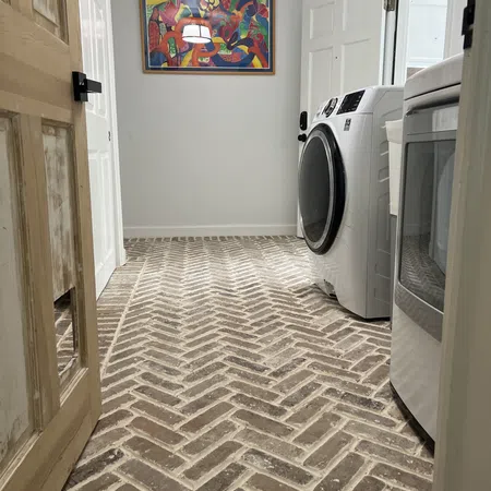 Laundry room with herringbone brick flooring, white washer and dryer, wooden door, and colorful abstract wall art.