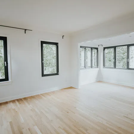 Bright empty room with light hardwood floors and large black-framed windows overlooking green trees.
