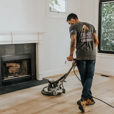 Worker using a floor sanding machine on hardwood floors in a bright room with a fireplace and large window.