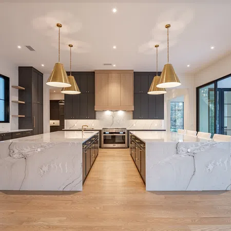 Modern kitchen with dual marble islands, gold pendant lights, dark cabinets, large windows, and open patio access.