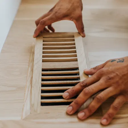 Hands installing a wooden vent cover on a light wood floor with a black wristwatch visible.
