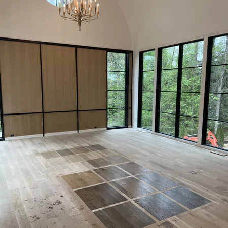 Modern bright room with large black-framed windows, wood floor samples, and an elegant chandelier under arched ceiling.