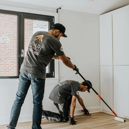 Two workers installing or finishing a hardwood floor in a bright, modern room with large window and white walls.