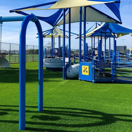 Modern playground with blue and beige shade sails on artificial grass under clear blue sky.