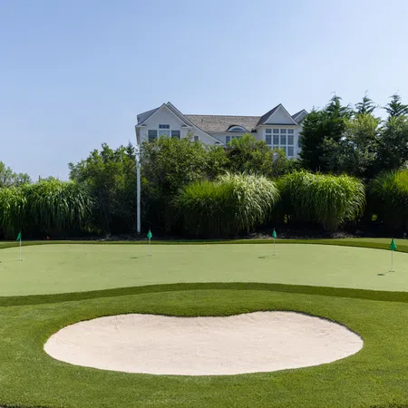 Golf putting green with flags, sand bunker in foreground, lush greenery, and house in background under clear sky