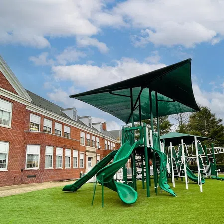 Green playground slides and swings set on artificial grass next to a large red brick school building under a blue sky.