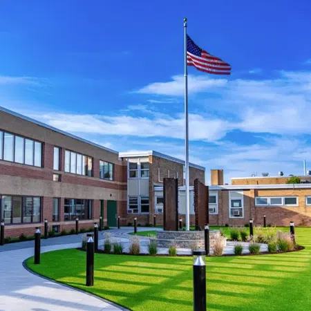 Modern school courtyard with green lawn, American flag on flagpole, and surrounding brick buildings under blue sky