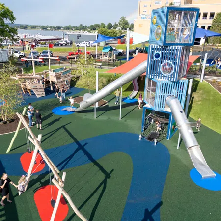 Modern playground with tall climbing tower, metal slides, swings, and colorful rubber flooring near waterfront park.