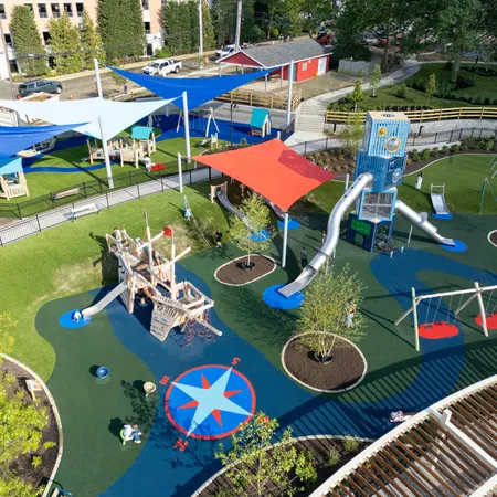 A colorful outdoor playground with slides, swings, shaded areas, climbing structures, and a compass design on the ground.